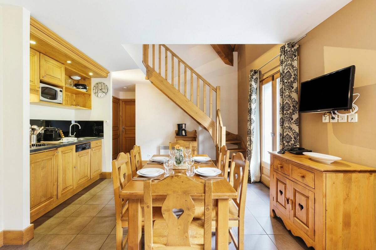 Une table à manger en bois avec des chaises, une cuisine avec des meubles en bois et un four à micro-ondes, un escalier et un téléviseur à écran plat fixé au mur.