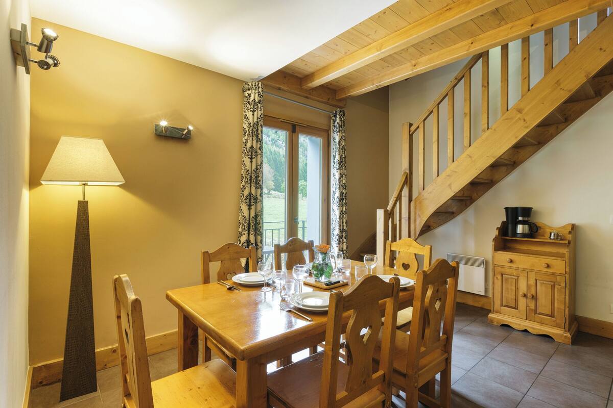 Un coin repas avec une table en bois dressée pour quatre personnes, une lampe et un escalier menant à l’étage supérieur.