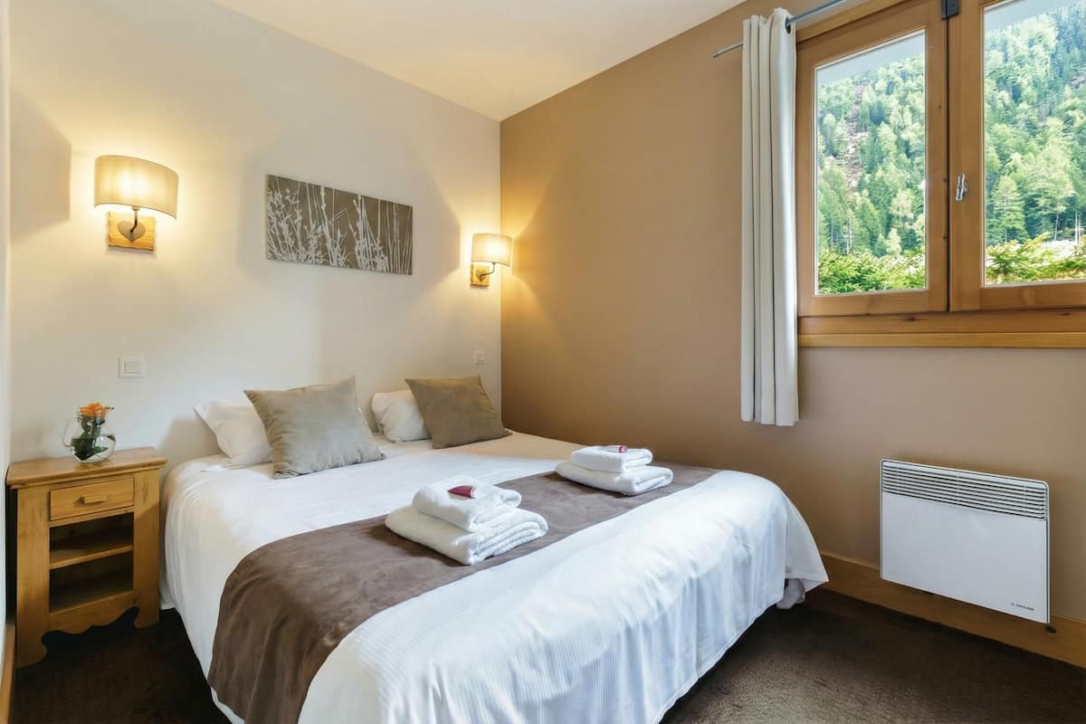 Une chambre à coucher comprenant un lit, des tables de chevet, des lampes fixées au mur, une fenêtre avec des rideaux et un radiateur.