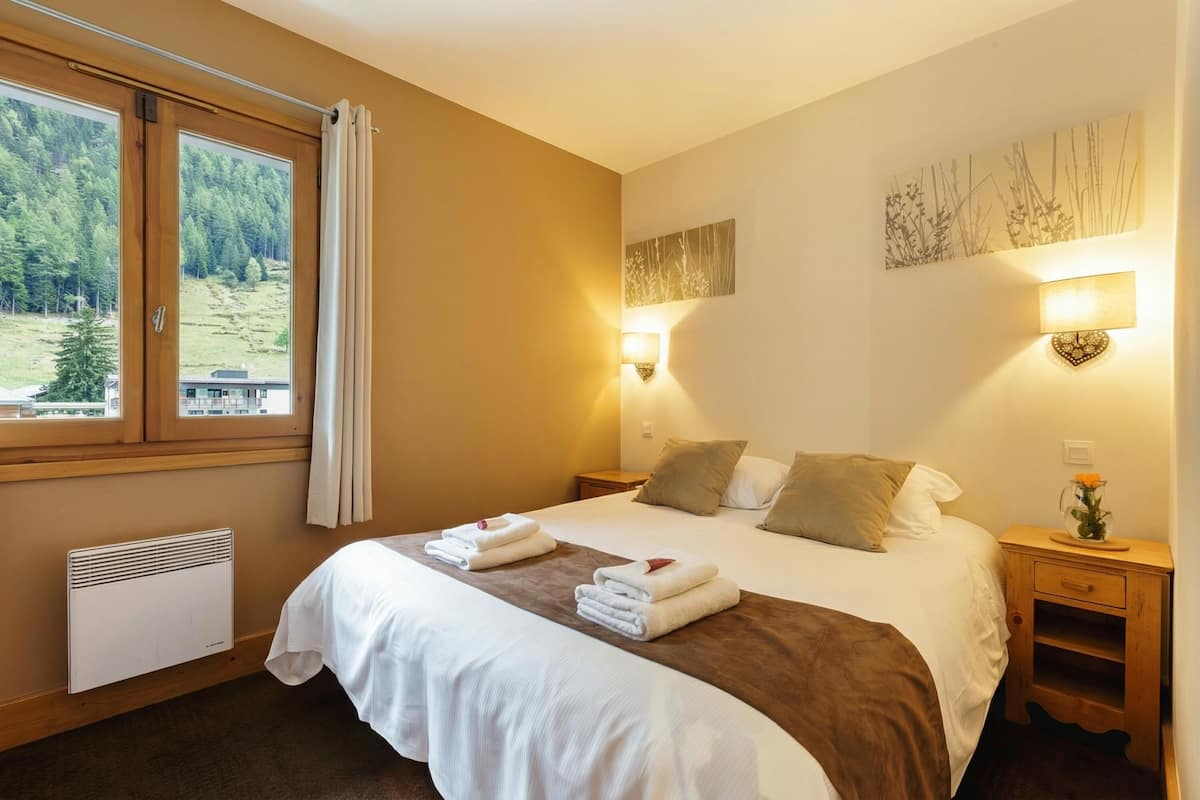 Une chambre d’hôtel avec un grand lit, deux tables de chevet, une lampe fixée au mur, un radiateur et une fenêtre avec des rideaux.