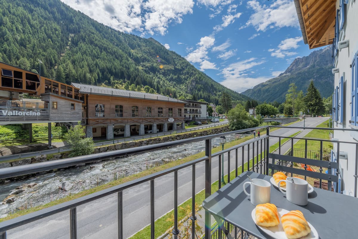 Un balcon avec une table préparée pour le petit-déjeuner, offrant une vue sur une rivière et des montagnes.
