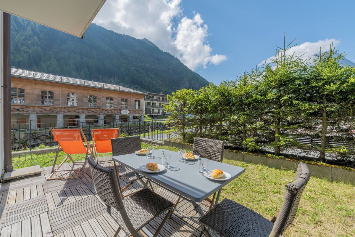 Un espace extérieur aménagé pour prendre les repas, avec une table dressée pour deux personnes, des chaises et une vue sur une montagne et un bâtiment en arrière-plan.