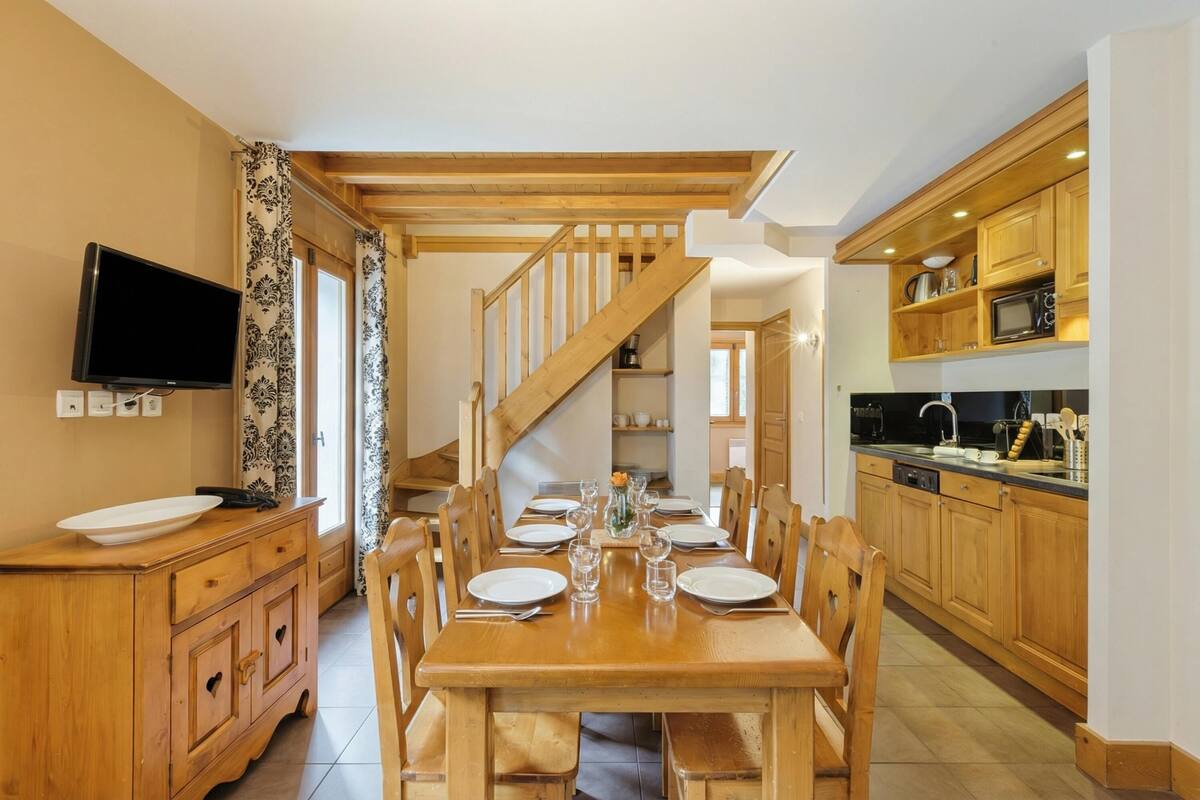 Un coin repas avec une table en bois dressée pour six personnes, bordé de meubles en bois et d’un téléviseur placé sur le côté gauche. Un escalier mène à l’étage supérieur.