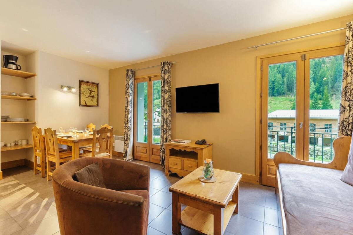Une pièce comprenant un canapé, une table en bois, un téléviseur, un coin repas avec des chaises, et offrant une vue sur la verdure grâce à de grandes fenêtres.