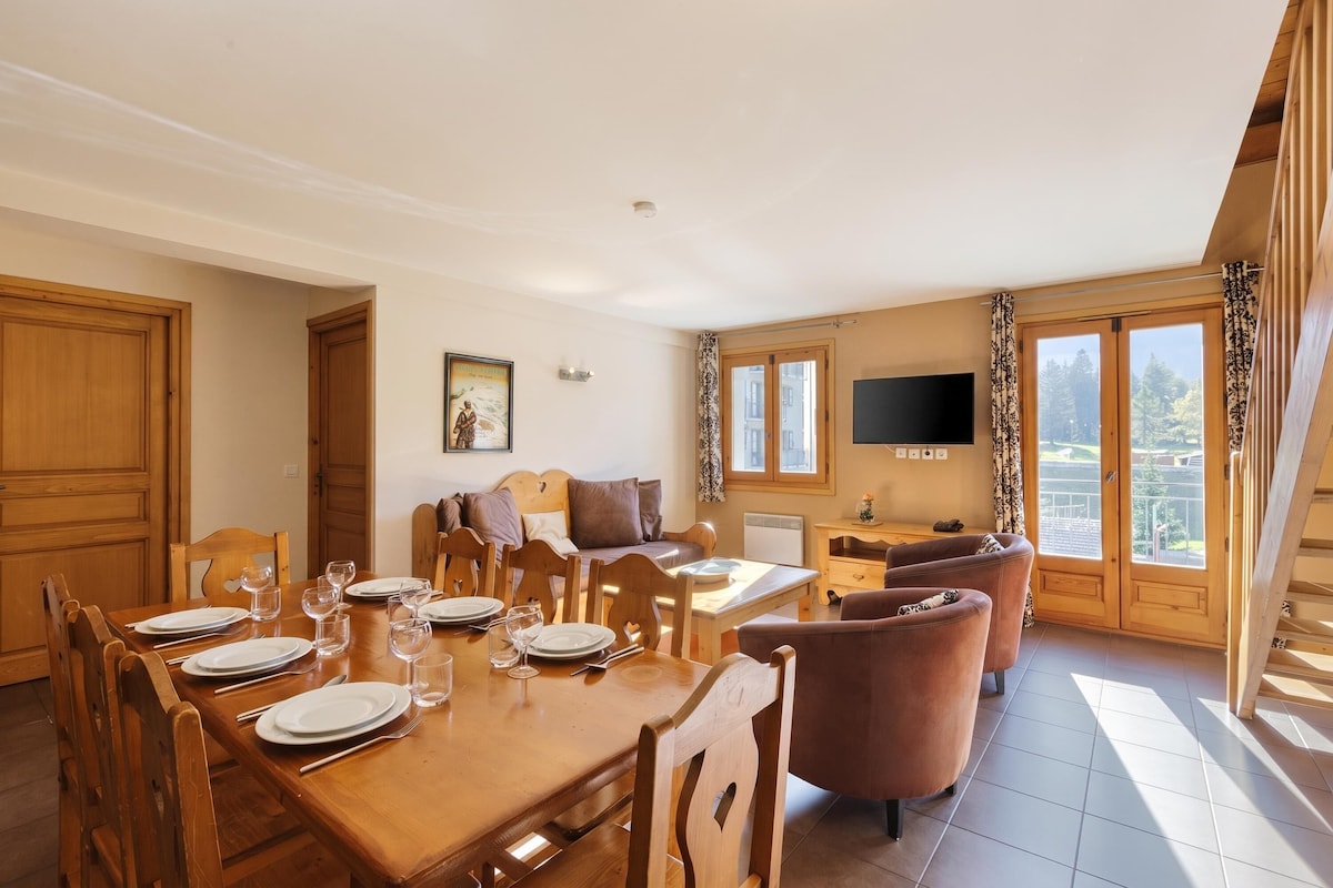 Un comedor con una mesa de madera dispuesta para una comida, un sofá y un televisor en una habitación con ventanas amplias.