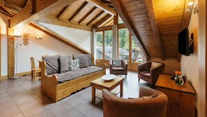 Ein gemütliches Loftzimmer mit Holzbalkendecke, einer Couch, einem Sessel, einem kleinen Tisch und einem an der Wand befestigten Fernseher.