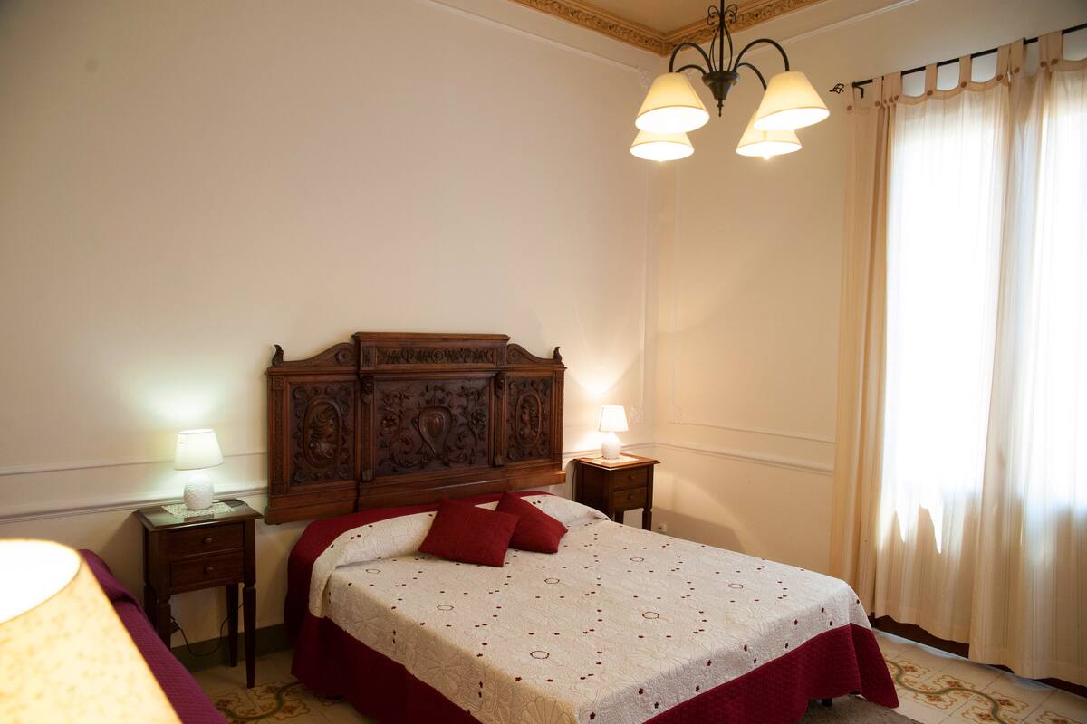 A bedroom with a wooden headboard, two bedside lamps, a bed with a red bedspread, and a window with curtains.