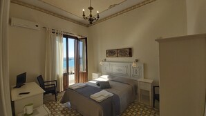 A bedroom with a bed, a desk, a chair, a nightstand, and a view of the sea through a window. - B&b Belveliero (Trapani)