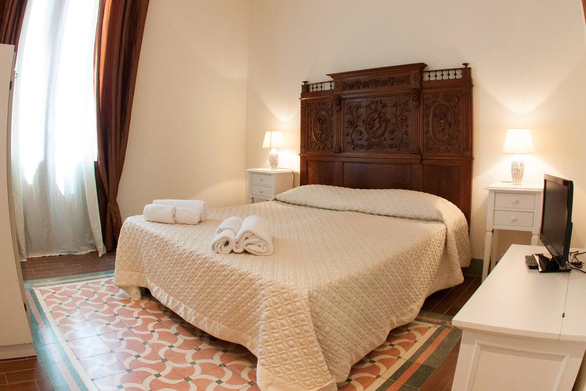 A neatly made bed with a quilt, two bedside tables with lamps, and a television on a desk.