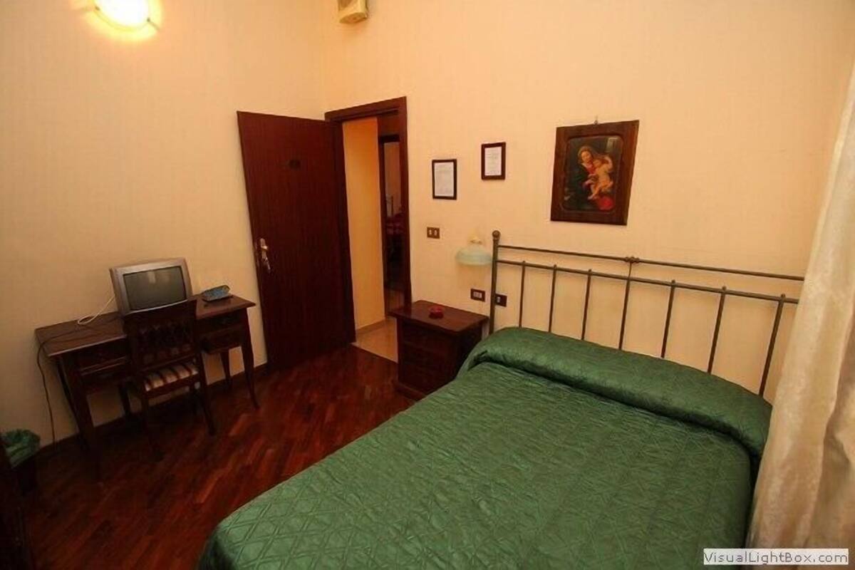 A bedroom with a bed, a desk with a computer, a chair, a door, and a wall-mounted picture.