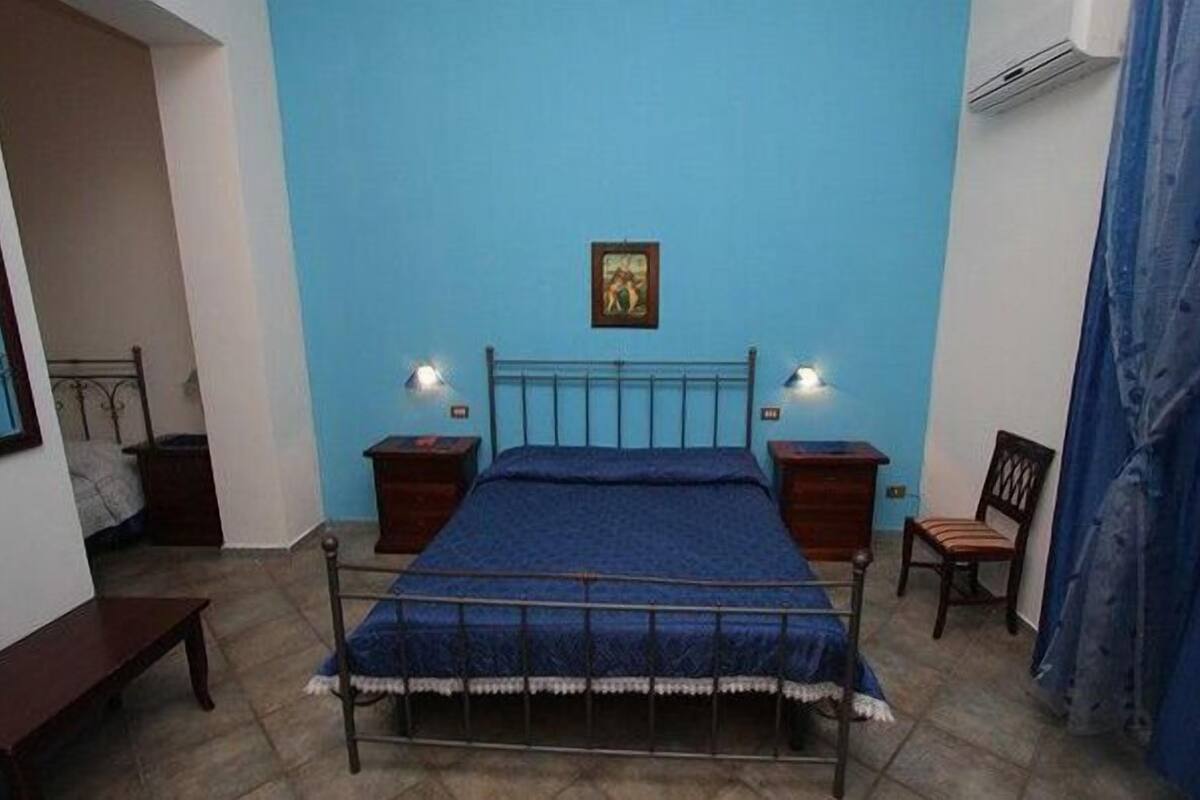A bedroom with a bed, two bedside tables, a chair, a picture on the wall, and an air conditioning unit.