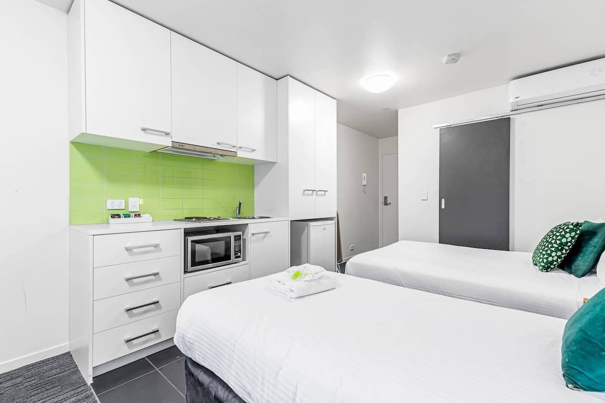 A modern hotel room with a kitchenette, a bed with green pillows, and a separate bathroom.