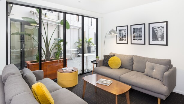 A modern living room with a grey sofa, a wooden coffee table, and a yellow ottoman. There are three framed pictures on the wall, and a large window with a view of the outside. - Quality Apartments North Melbourne (North Melbourne)