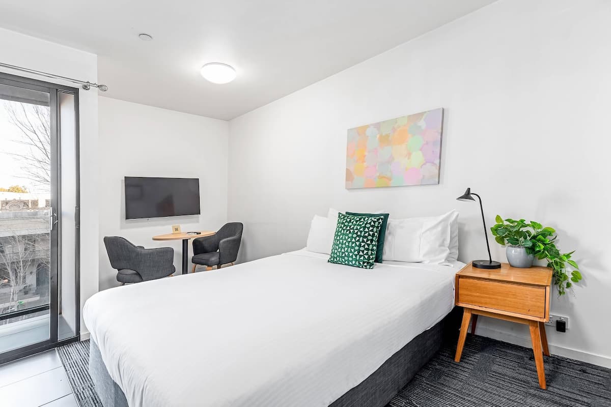 A hotel room with a bed, a TV, a desk with chairs, a plant, and a window with a view.