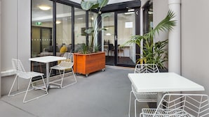 An outdoor seating area with white tables and chairs, surrounded by potted plants and a wooden planter. - Quality Apartments North Melbourne (North Melbourne)