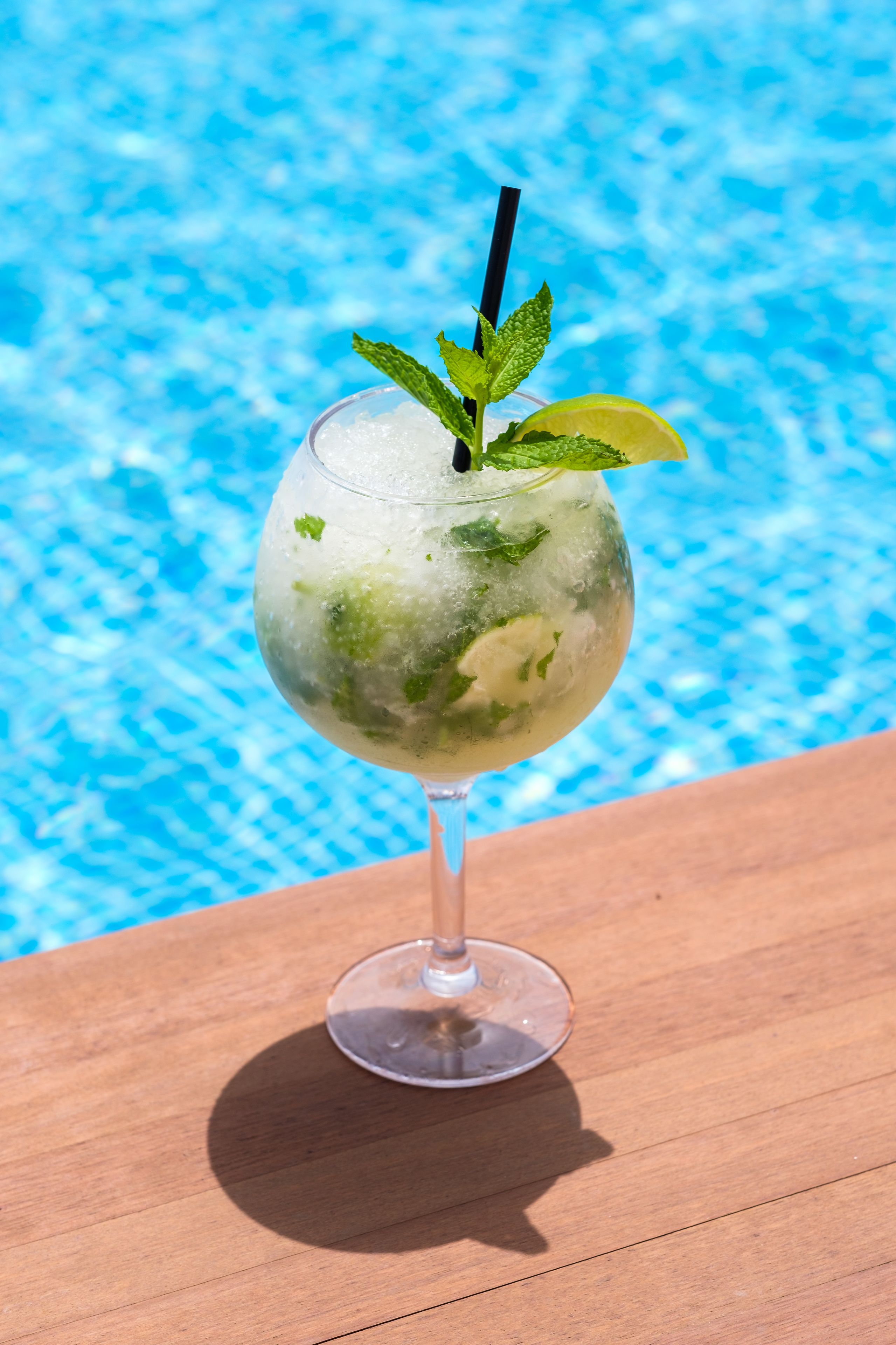 A cocktail with mint leaves and a lime wedge, placed on a wooden surface by a pool.