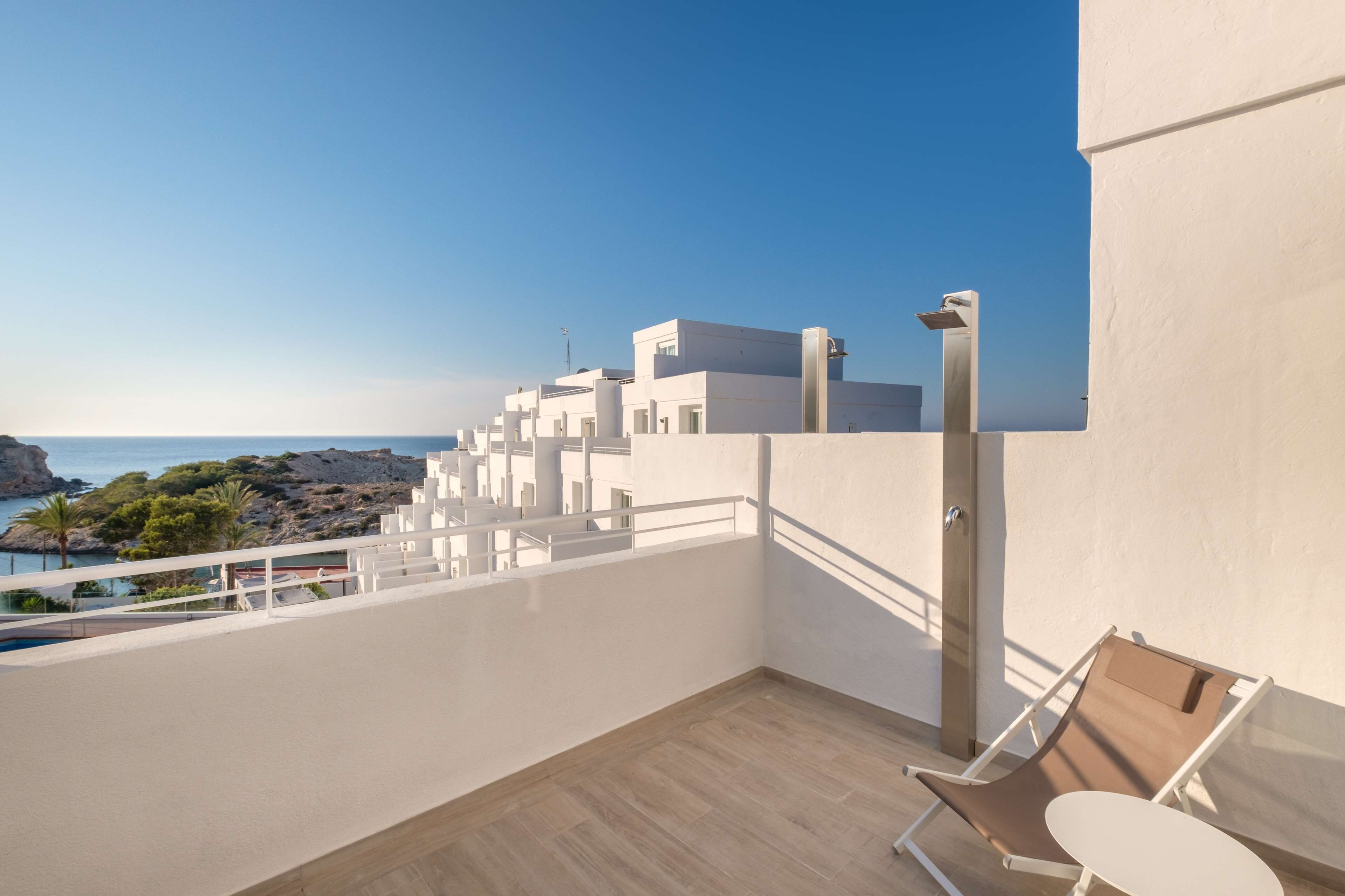 A balcony with a view of the sea and rocky landscape, a white railing, and a pair of chairs and a table.