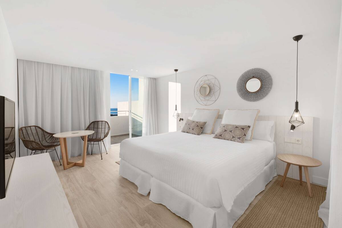 Ein modernes Hotelzimmer mit einem großen Bett, einem Balkon mit Vorhängen, einer Essecke mit Stühlen und einem kleinen Tisch.