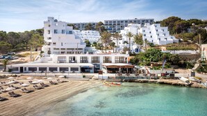 Un hôtel situé sur la côte, composé de plusieurs bâtiments blancs, avec une plage dotée de transats et offrant une vue dégagée sur la mer.