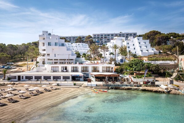 A coastal hotel with multiple white buildings, a beach with sun loungers, and a clear view of the sea.