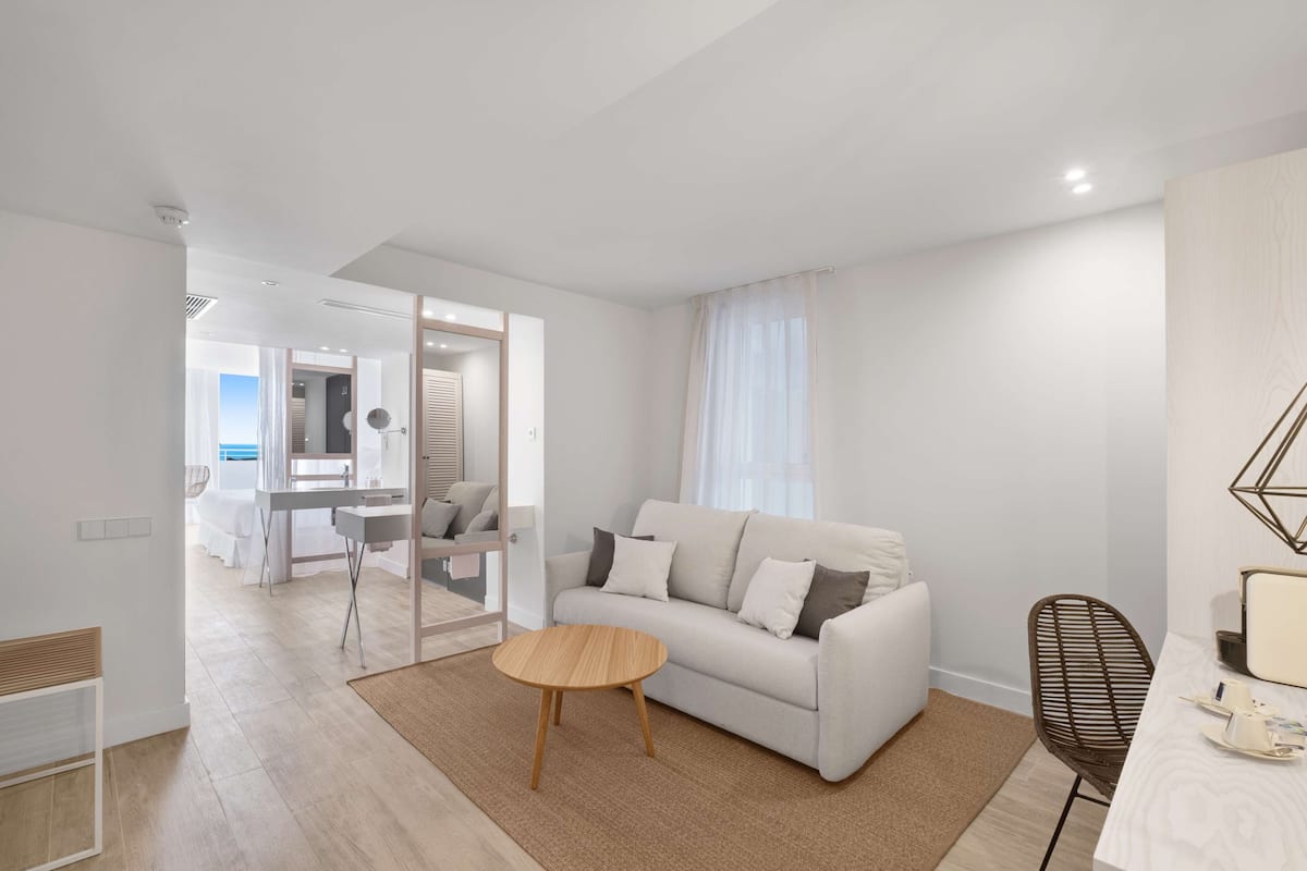 Ein modernes Wohnzimmer mit einer Couch, einem runden Couchtisch und einem Sessel. Im Hintergrund steht ein Schreibtisch mit Stuhl und ein Bett.