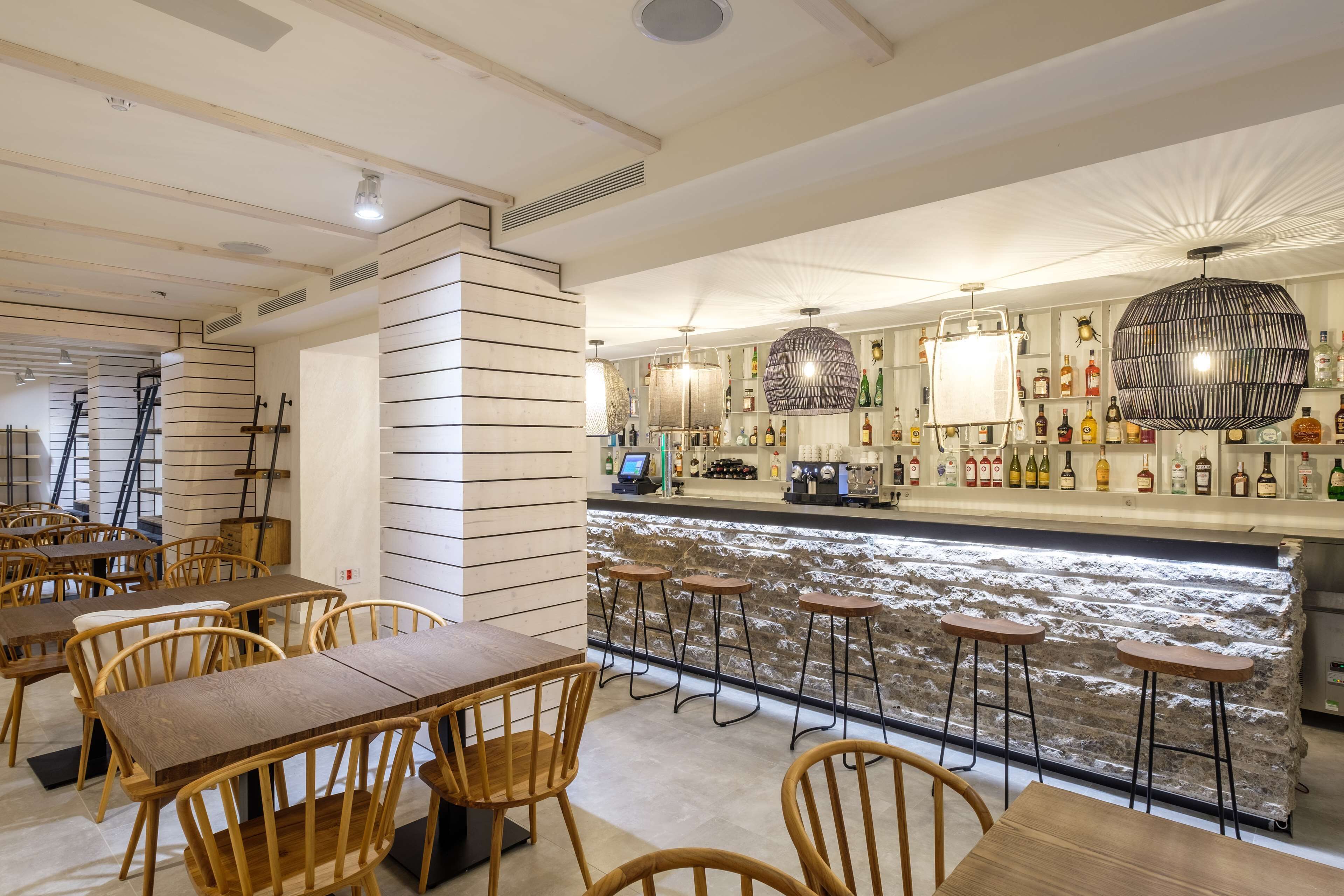 Un área de bar con mesas y taburetes de madera, una barra de piedra y estantes con botellas.