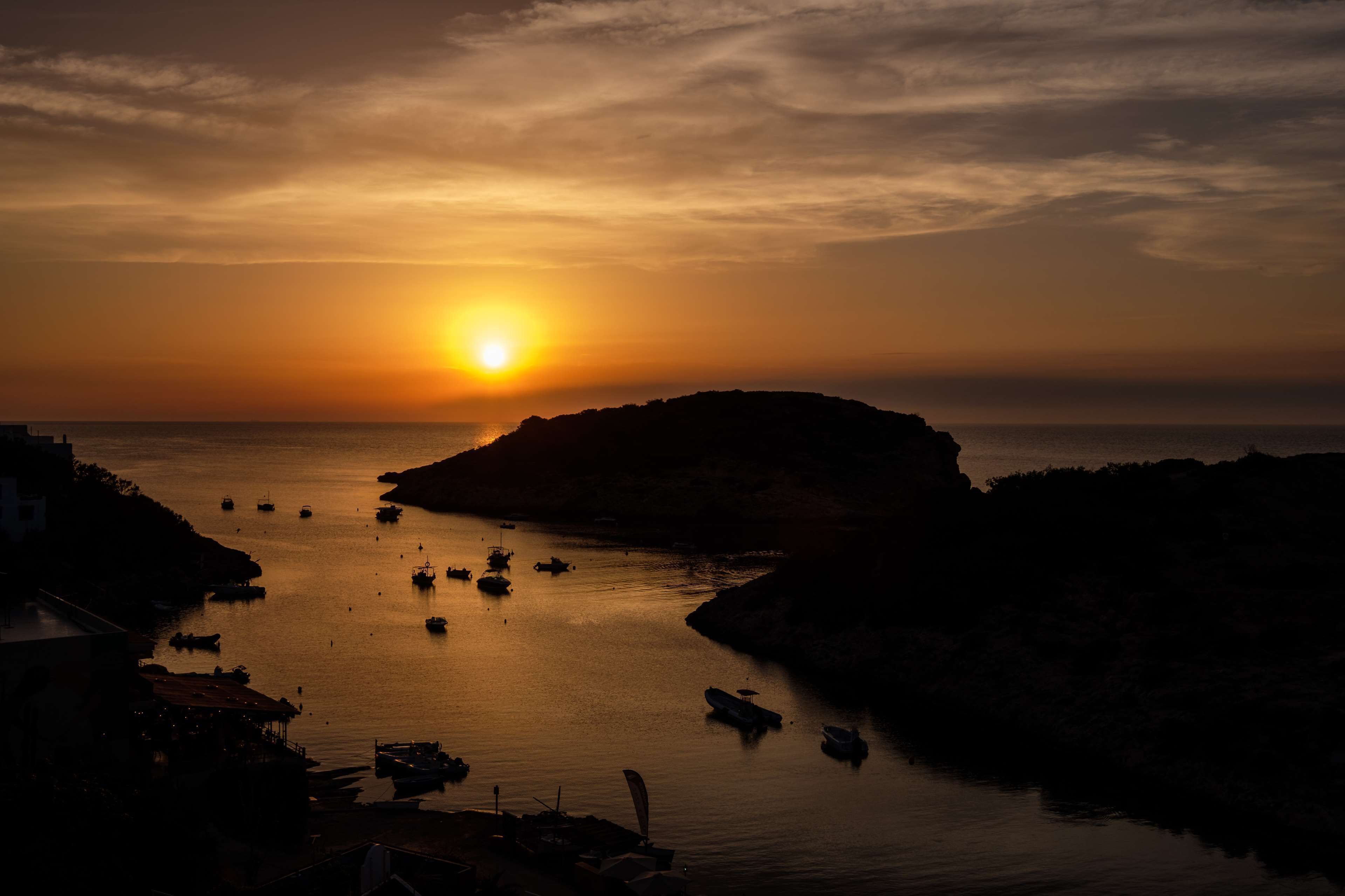 Vista de un atardecer costero con barcos y terreno rocoso.