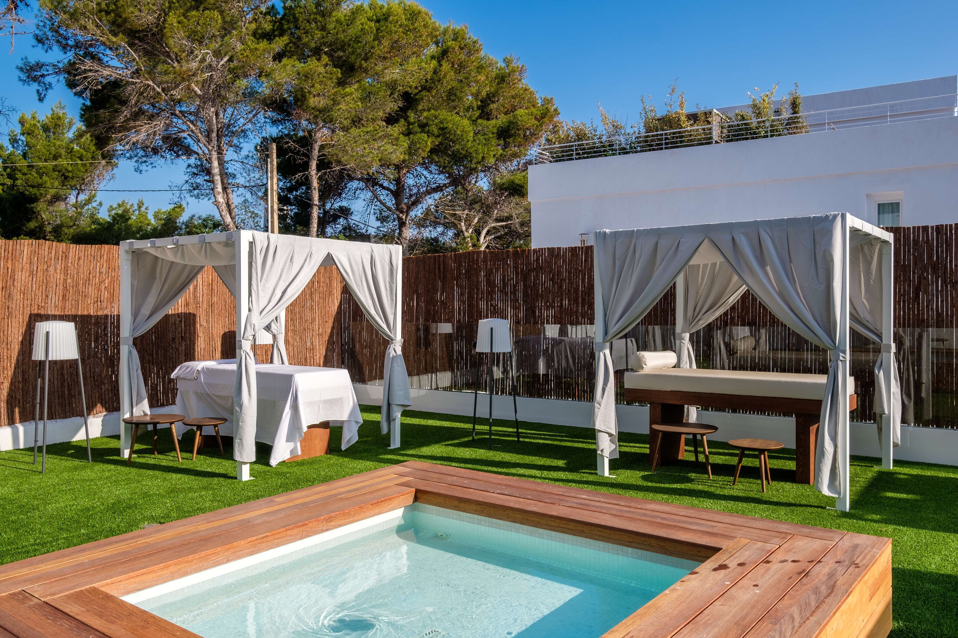 Un espace piscine extérieur moderne, doté d’une terrasse en bois, de deux auvents blancs et d’un bâtiment blanc en arrière-plan.