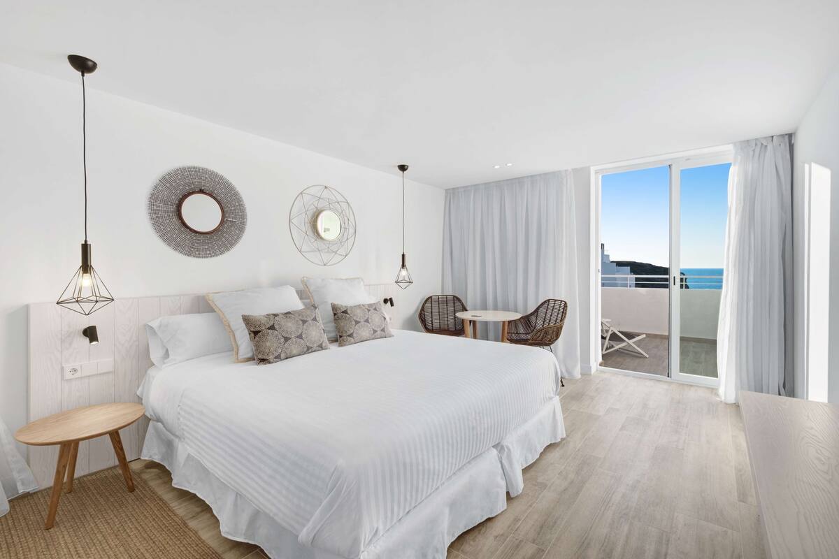 Ein modernes Schlafzimmer mit einem großen Bett, zwei Wandleuchten, einem runden Spiegel und Blick auf das Meer durch eine Schiebeglastür.