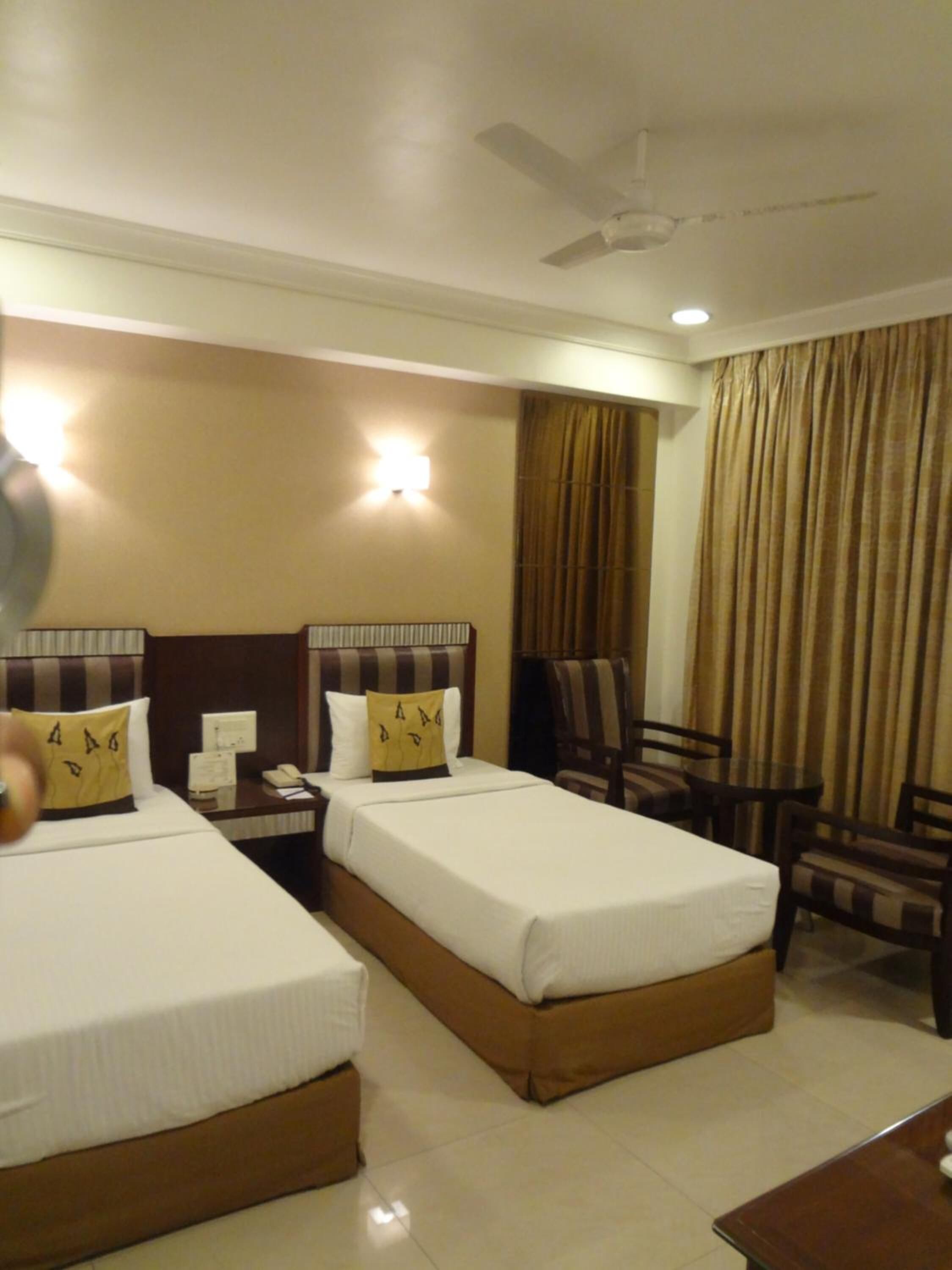 Quarto de hotel com duas camas, uma escrivaninha, uma cadeira, uma mesa, um ventilador de teto e uma luminária na parede.