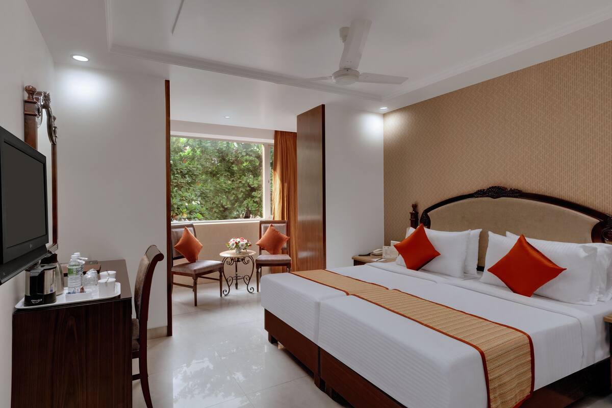 A hotel room with a large bed, a desk with a TV, a small table with a vase, and a window with curtains.