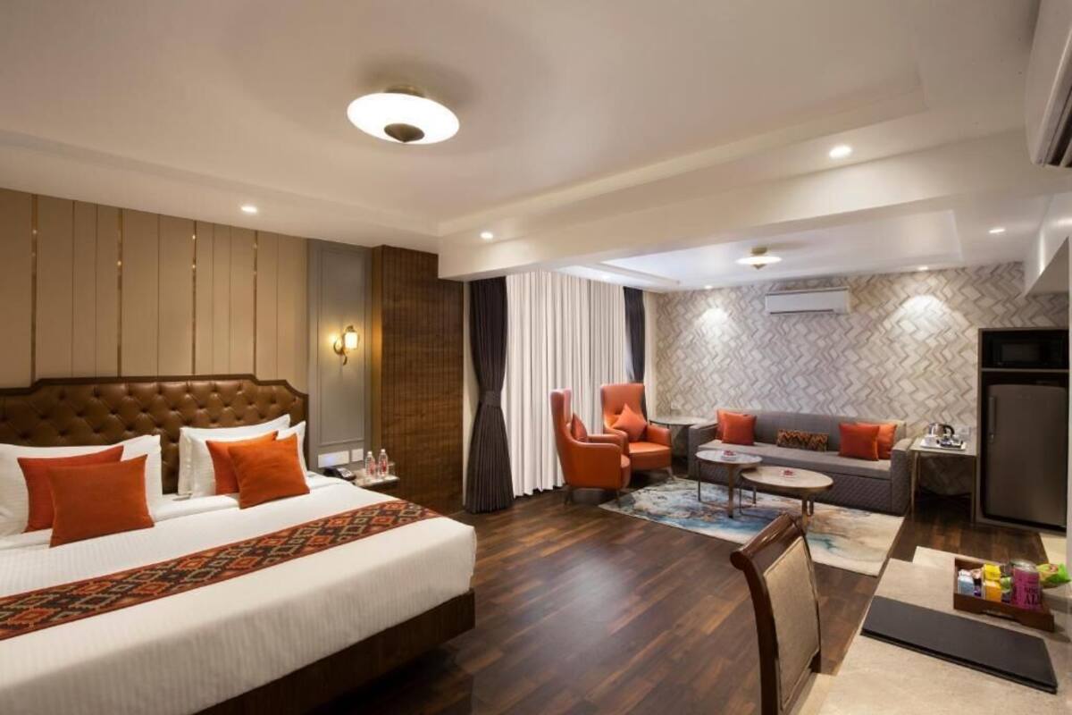 A hotel room with a large bed, a sofa, a chair, a small table, and an air conditioning unit.