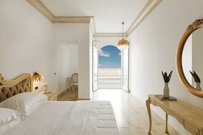 A bedroom with a large bed, a nightstand with a lamp, a dresser, and a mirror. There is a balcony with a view of the sky.