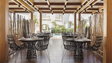 A covered outdoor dining area with multiple tables and chairs, a wooden ceiling, and a view of a pool and building in the background.