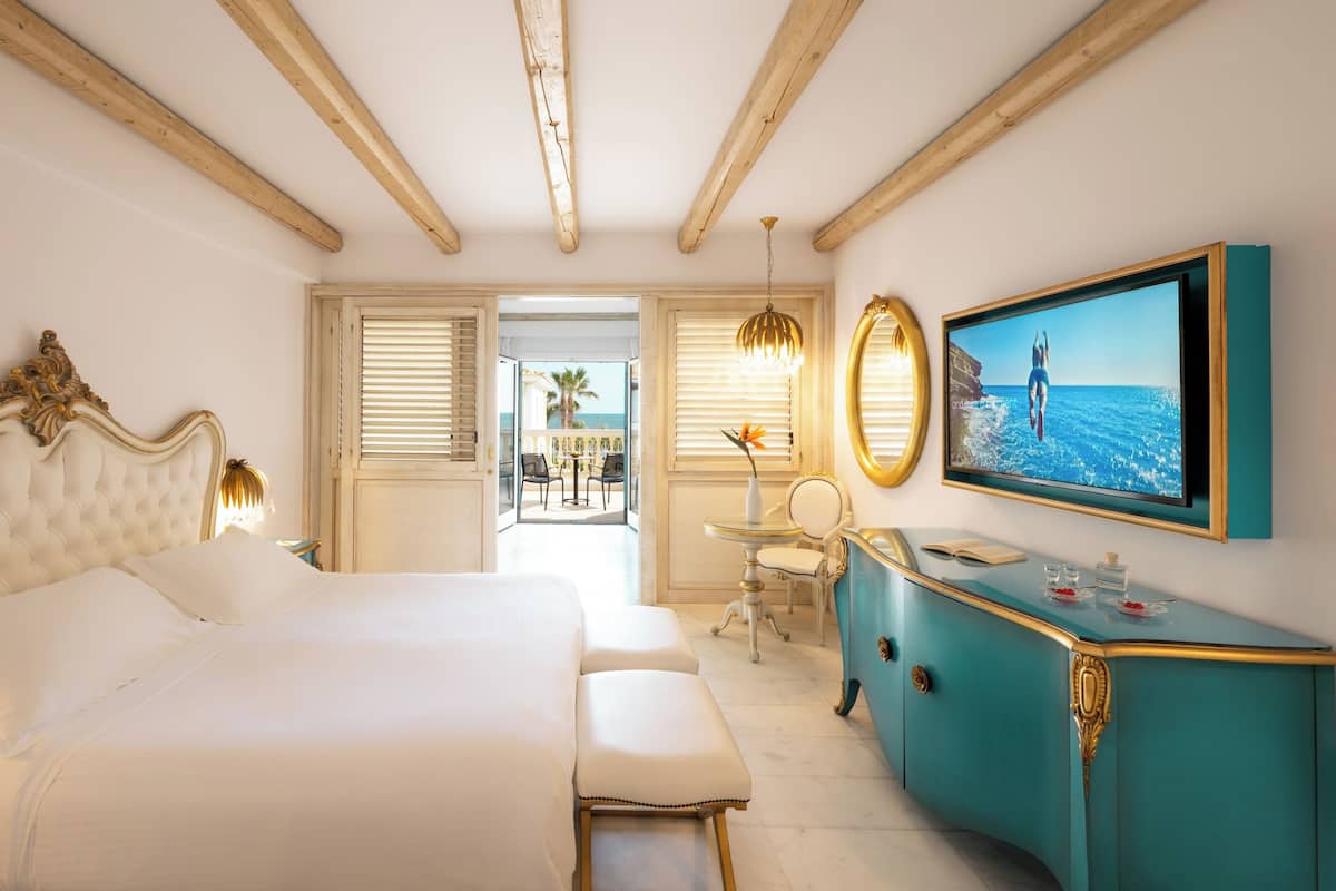 Un dormitorio con cama, escritorio, silla, cómoda, televisor y balcón con vista al mar.