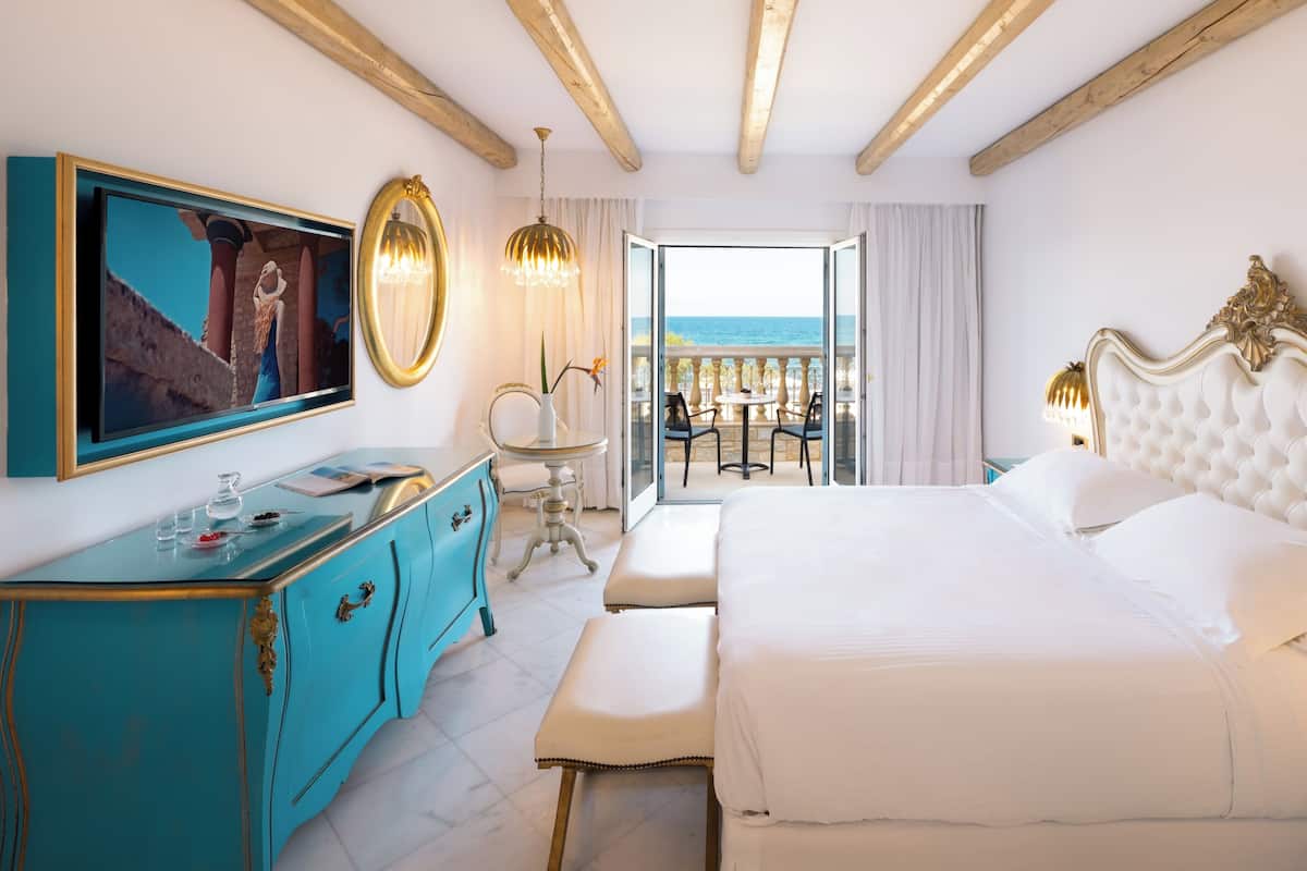 Una habitación de hotel con cama, televisor, cómoda, balcón con vistas y candelabro.