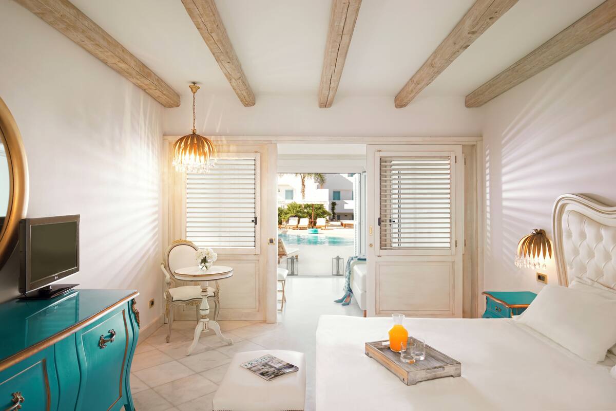 Un dormitorio con cama, televisor en una cómoda, mesita de noche con vaso de jugo de naranja y vista a una piscina y mobiliario exterior.