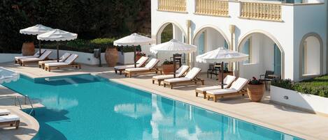 A pool area with lounge chairs and umbrellas, a clear pool, and a building with arched windows and balconies.