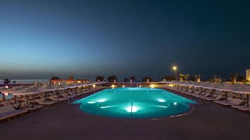 Zona junto a la piscina con sillones de playa y sombrillas, piscina y vista al mar al atardecer.