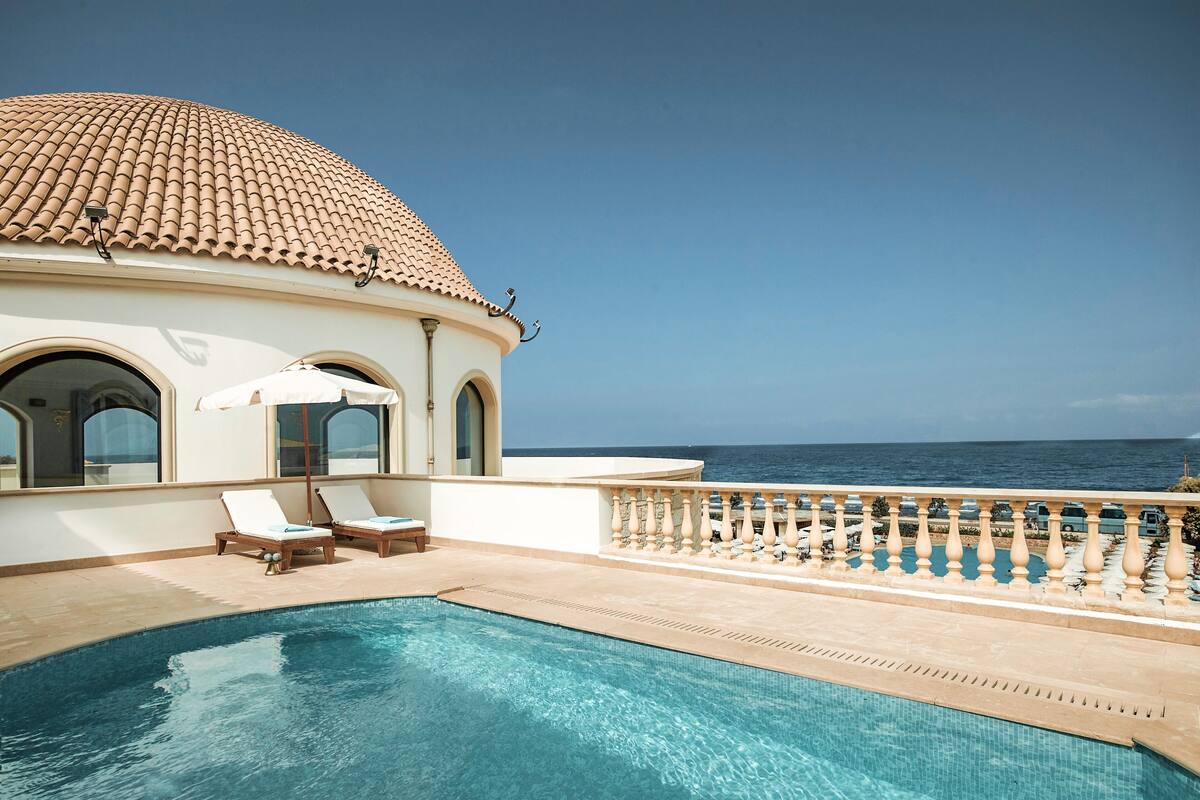 Un área de piscina con vistas despejadas al mar, un edificio blanco con techo de tejas y dos sillas de descanso.