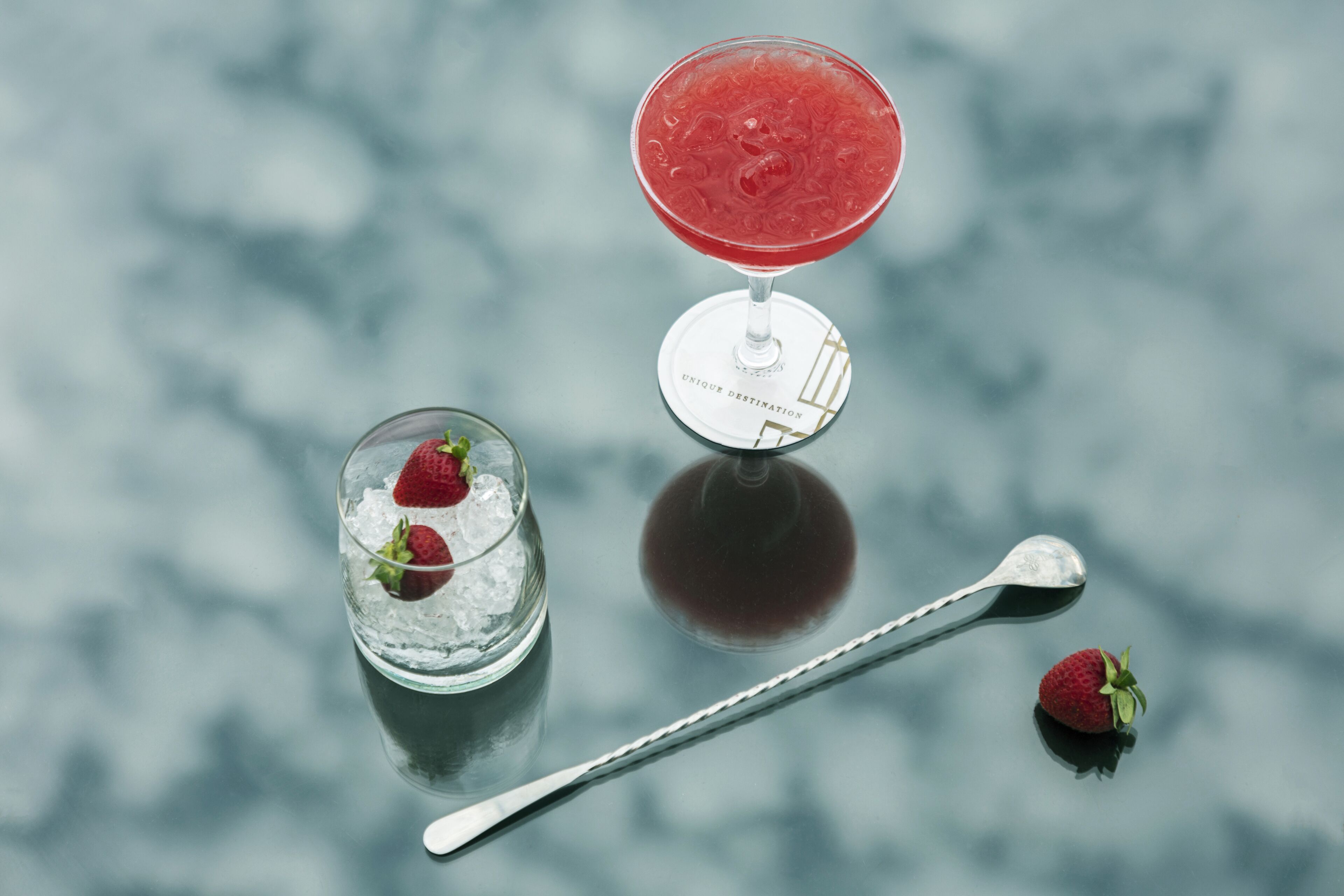 A martini glass with a red drink, a small glass with ice and strawberries, and a spoon on a reflective surface.