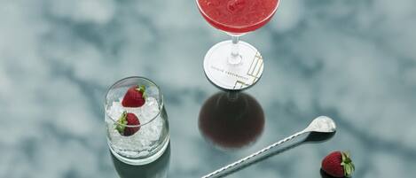 Un verre à martini contenant une boisson rouge, un petit verre avec des glaçons et des fraises, et une cuillère posée sur une surface réfléchissante.