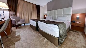 A hotel room with two beds, a desk, a chair, and a nightstand with a lamp.
