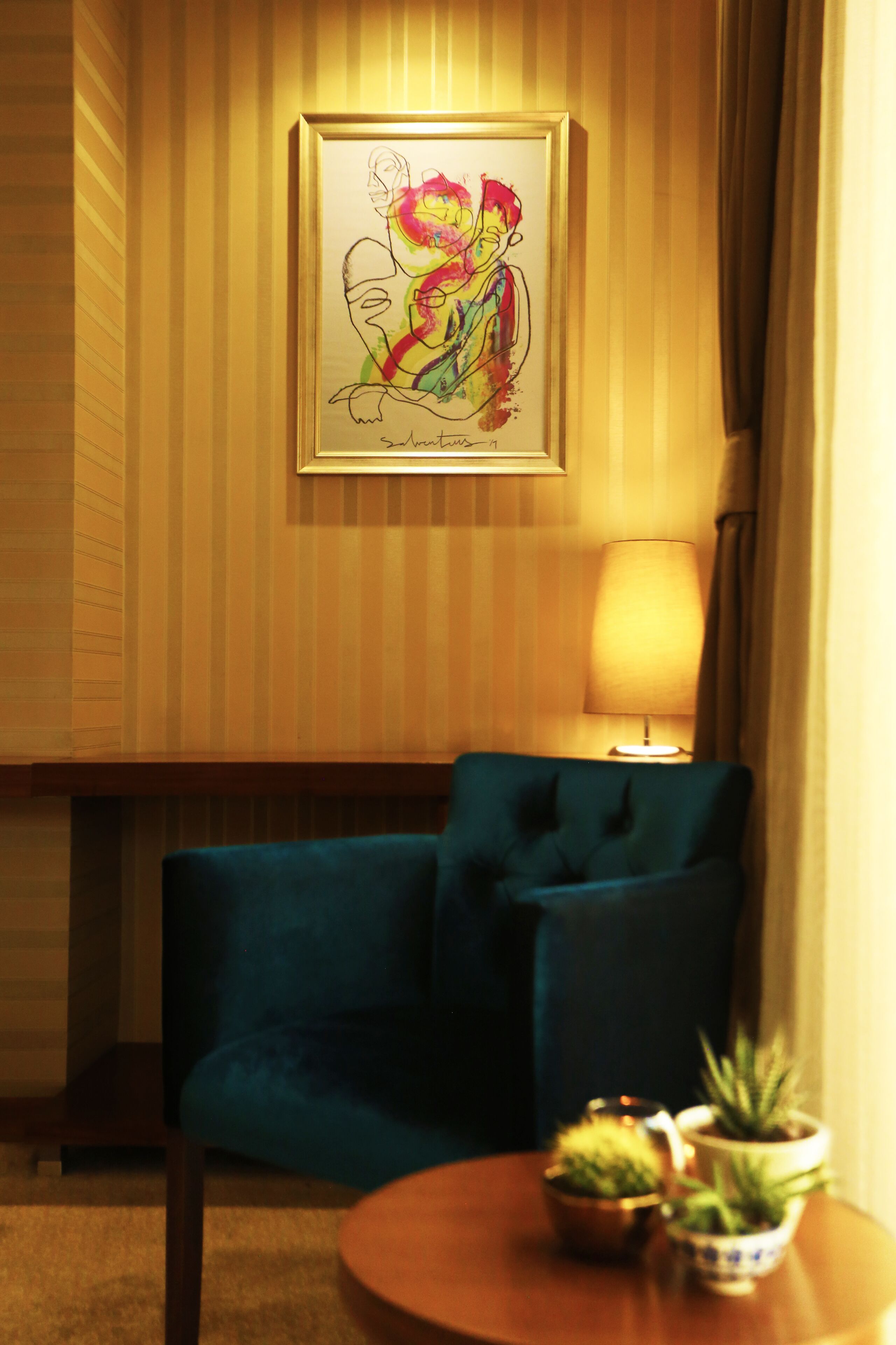 A room with a framed artwork on the wall, a blue armchair, a round wooden table with potted plants, and a lamp.