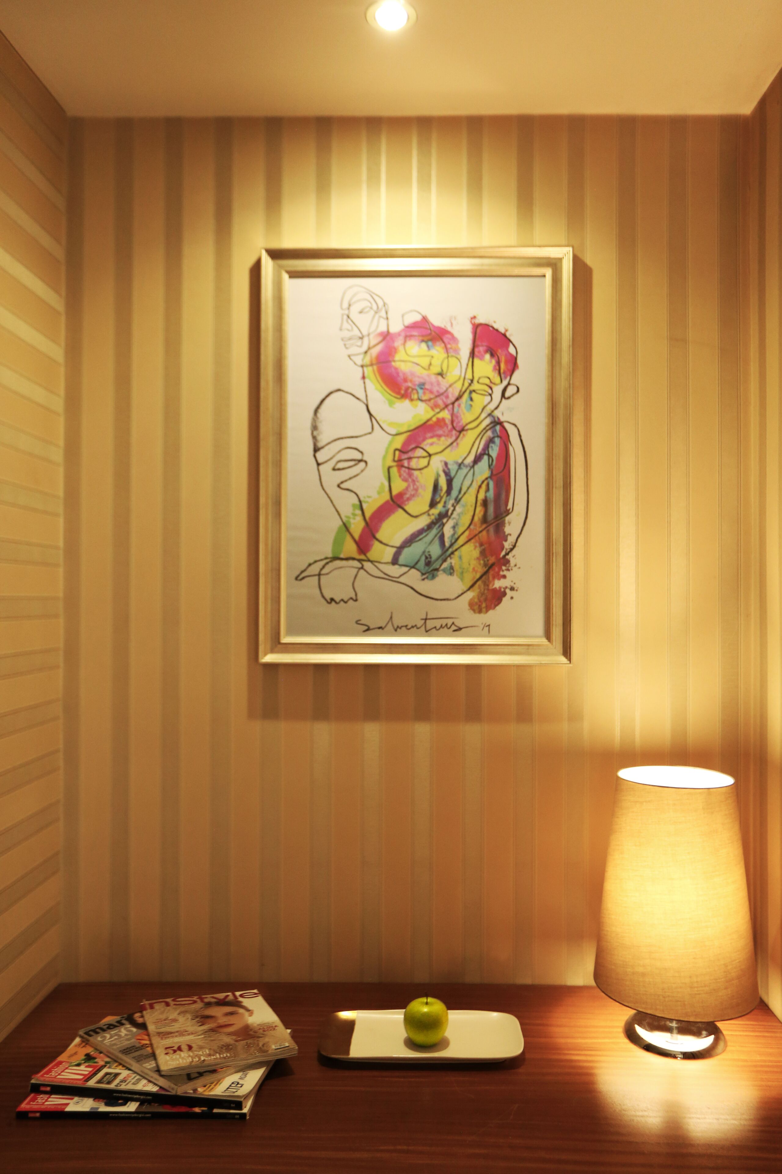 Un cuadro abstracto enmarcado en una pared, una mesa con revistas y una manzana, y una lámpara.