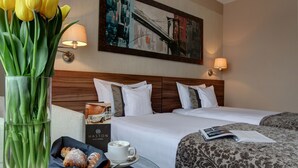A hotel room with two beds, a framed cityscape painting, a vase with yellow tulips, a basket of croissants, and a cup of coffee. - Haston City Hotel (Wroclaw)