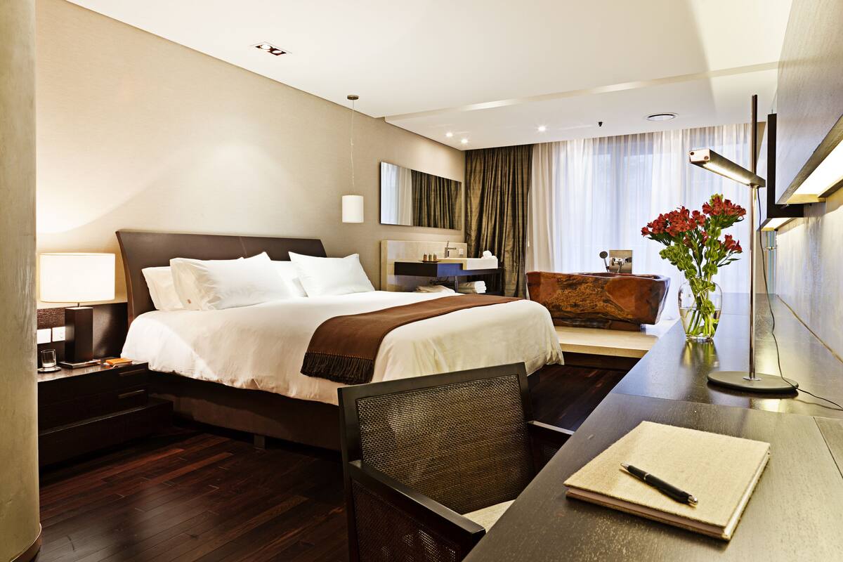 A modern hotel room with a large bed, bedside table, lamp, and a desk with a chair and a vase of flowers.