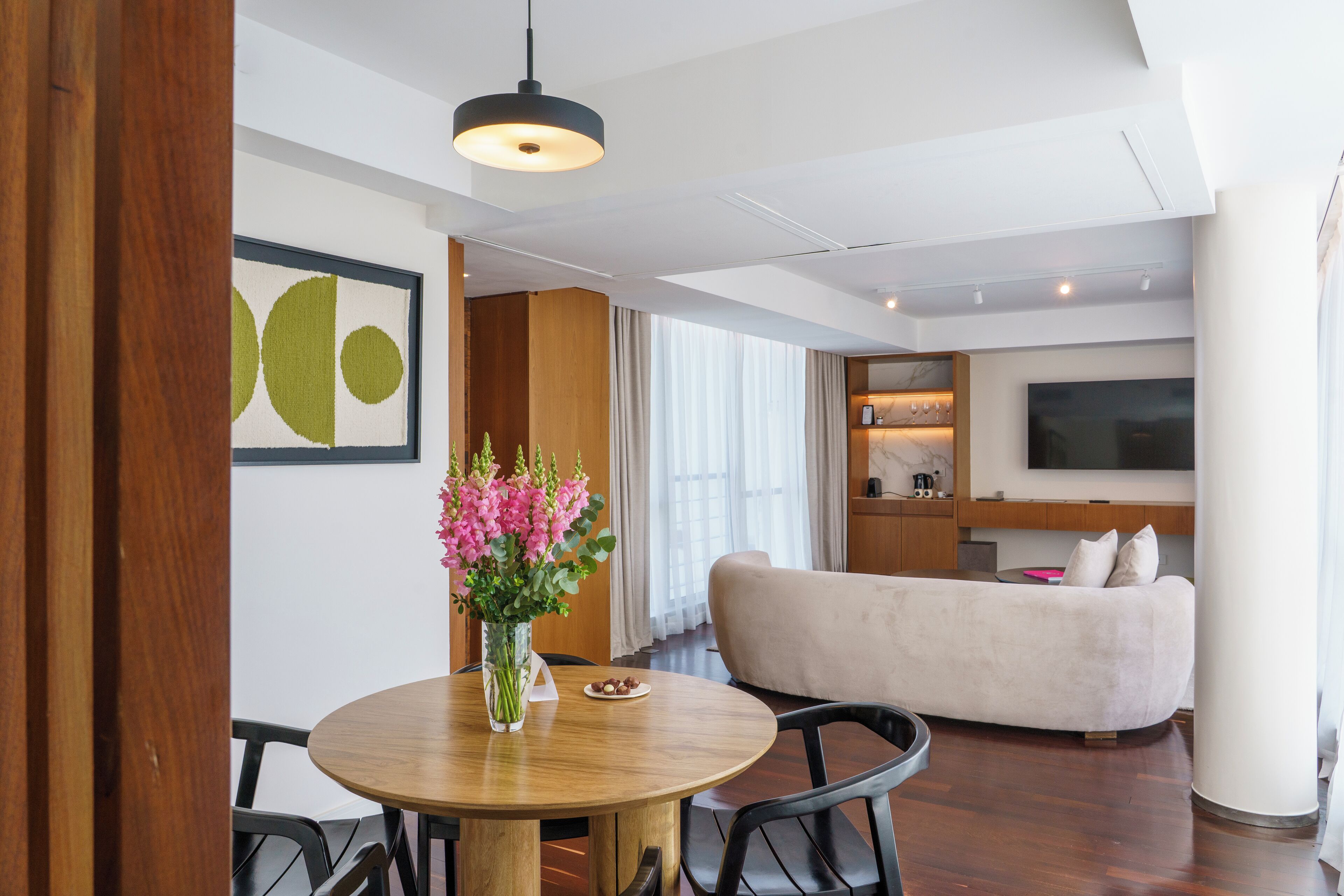 A modern living room with a round wooden table, a beige sofa, and a flat-screen TV mounted on the wall.