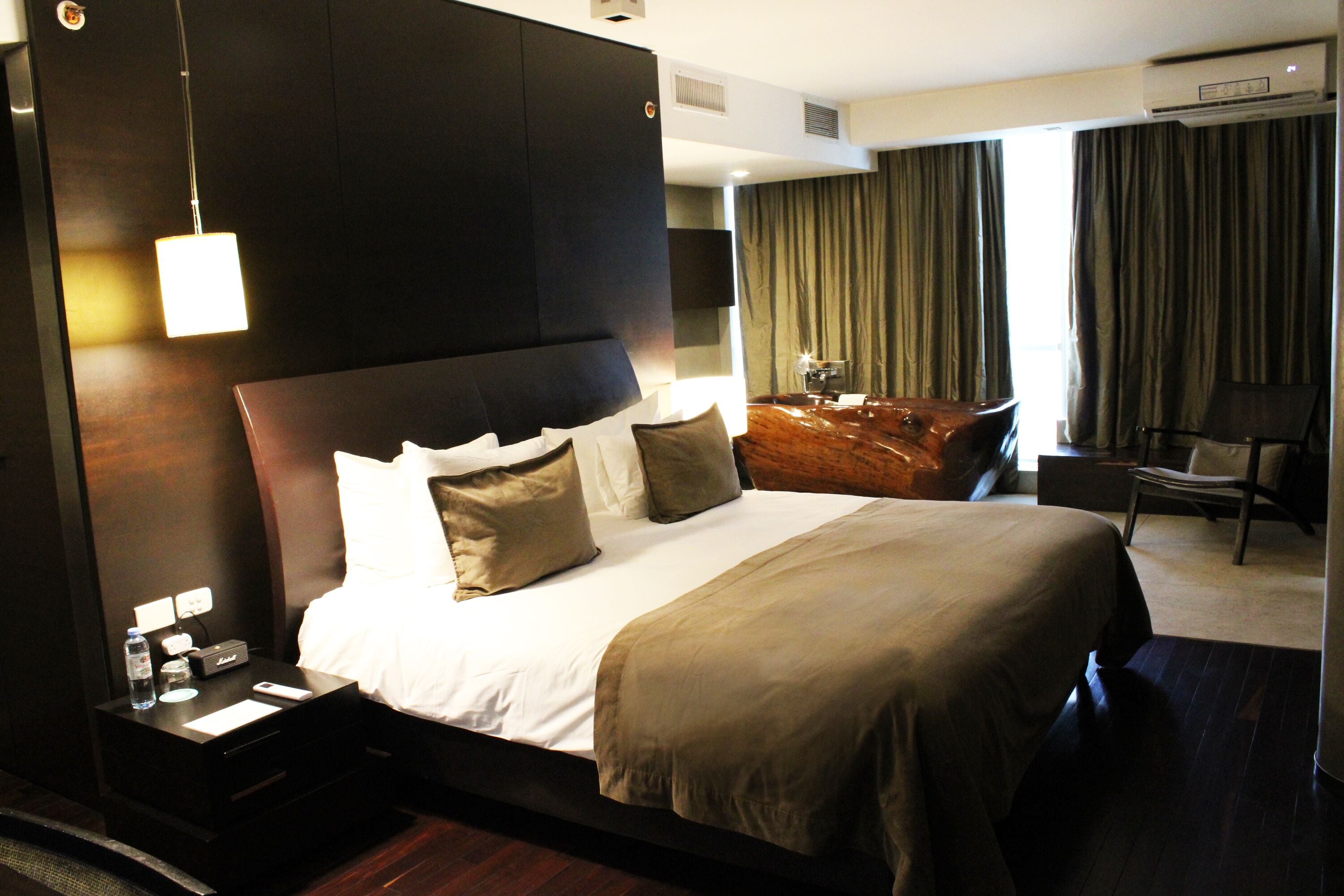 A hotel room with a large bed, a nightstand with a water bottle, a remote control, and a wooden side table with a lamp.