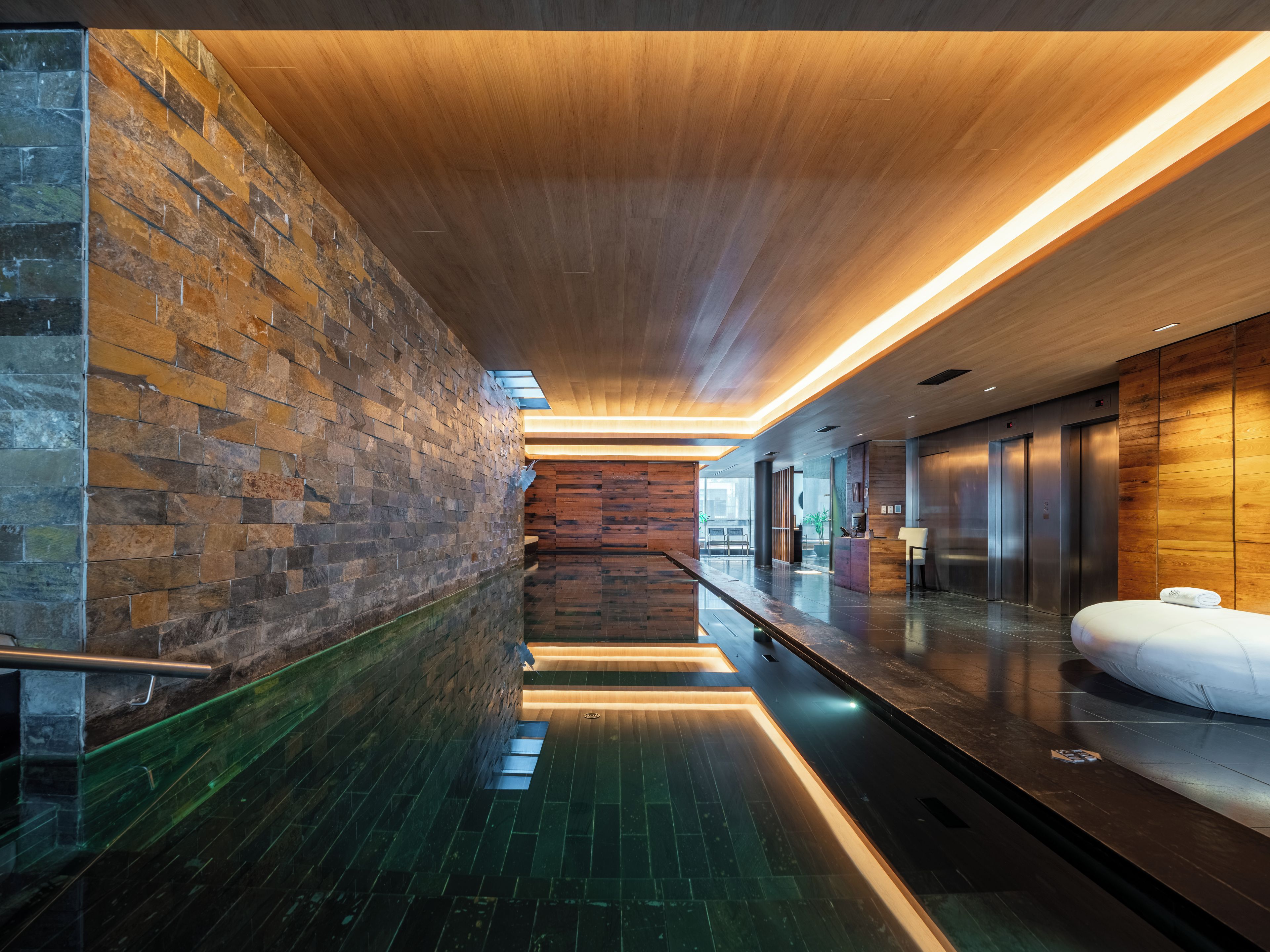 Un pasillo moderno de hotel con techo de madera, pared de piedra y barandilla de vidrio.
